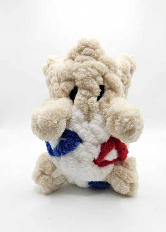 Togepi bag charm hanging half out of a white shell with blue and red triangle details embroidered into it in chenille yarn.